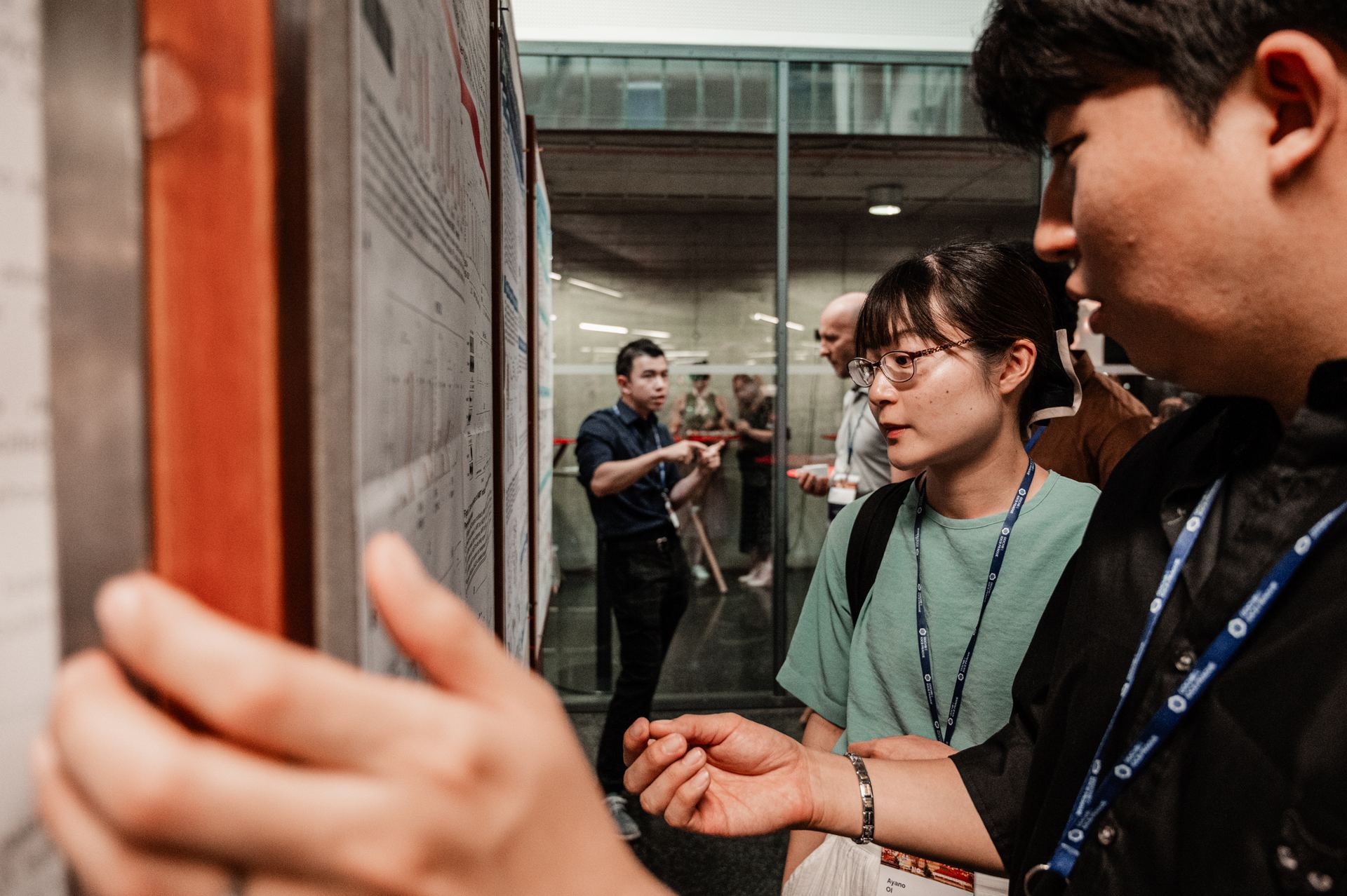 Two researchers listening as a third explains a poster, hand raised mid-gesture.